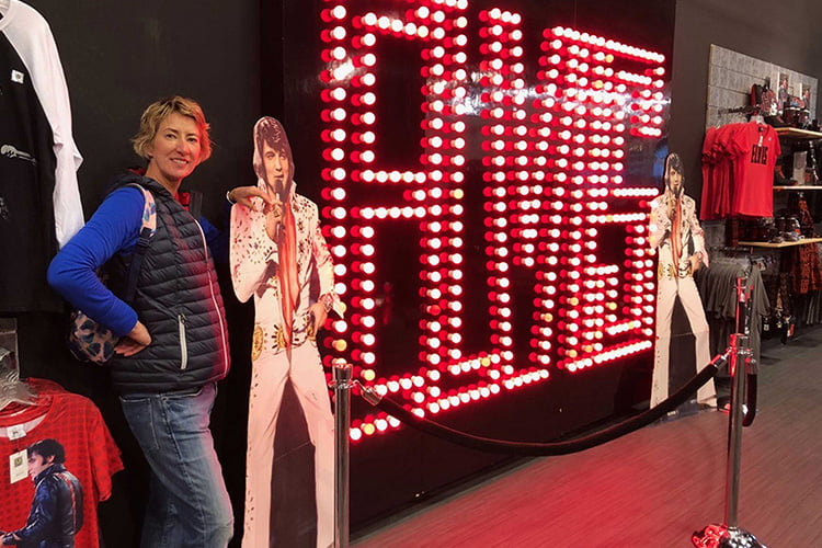 A blonde woman leaning with her arm on the shoulder of an Elvis Presley life size cutout with a large illuminated letter sign saying Elvis in yellow and red lightbulbs