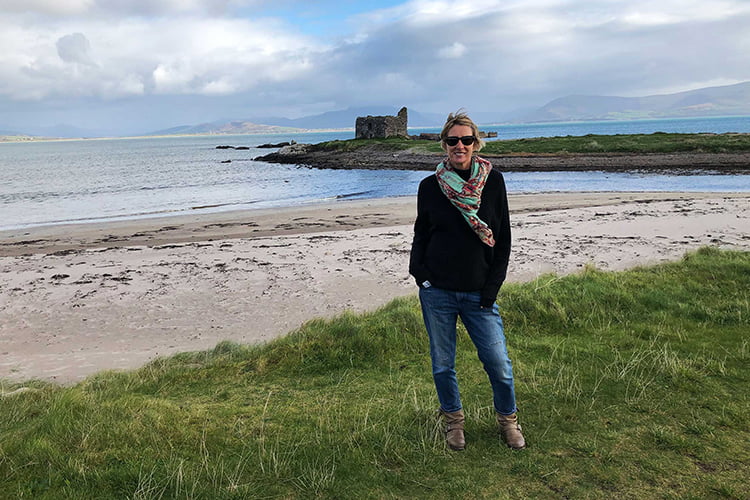 A blonde woman in black jumper and sunglasses standing on a deserted remote beach in front of a ruined building in The Ring of Kerry, Ireland