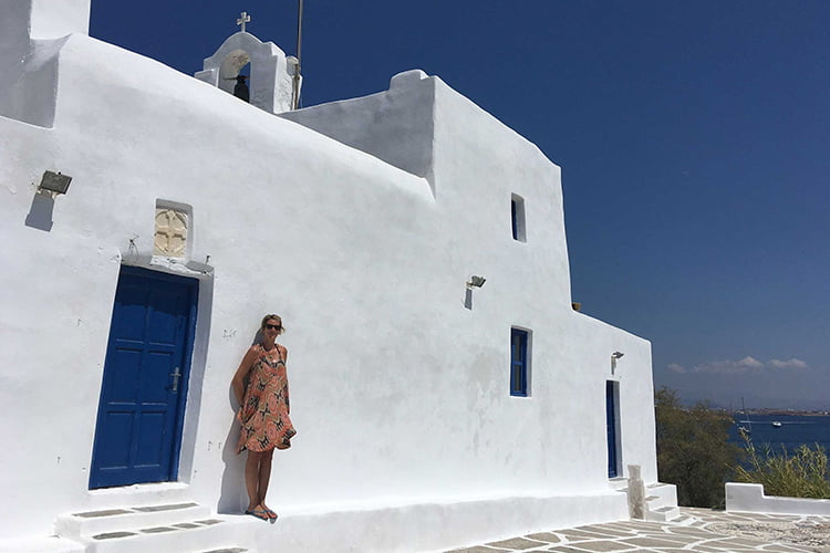 A woman in a patterned dress leans up a white wall outside a church, Milos Greece