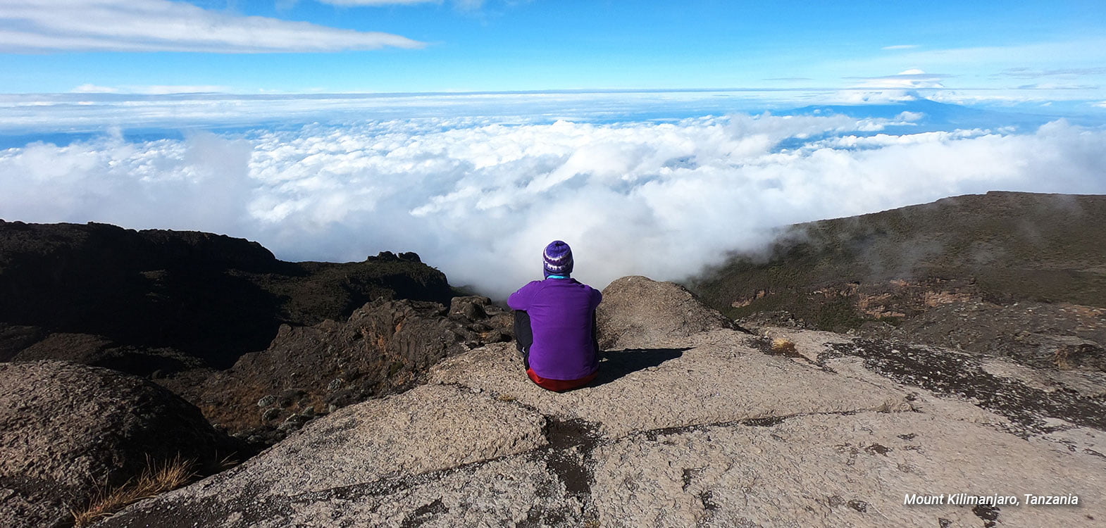 A blonde woman in a purple top and wooly hat sitting on a large flat rock high above the clouds at the top of a mountain