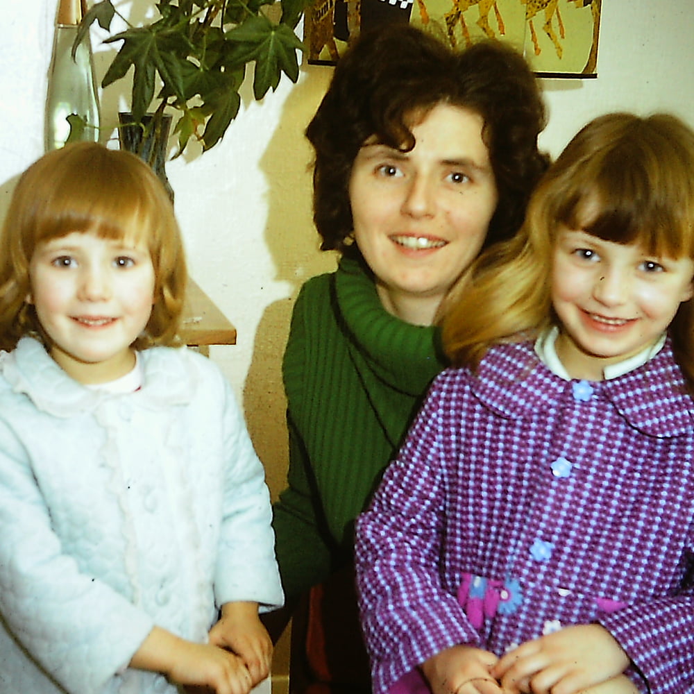 A dark haired woman in a green jumper and two young children in dressing gowns stand smiling at the camera