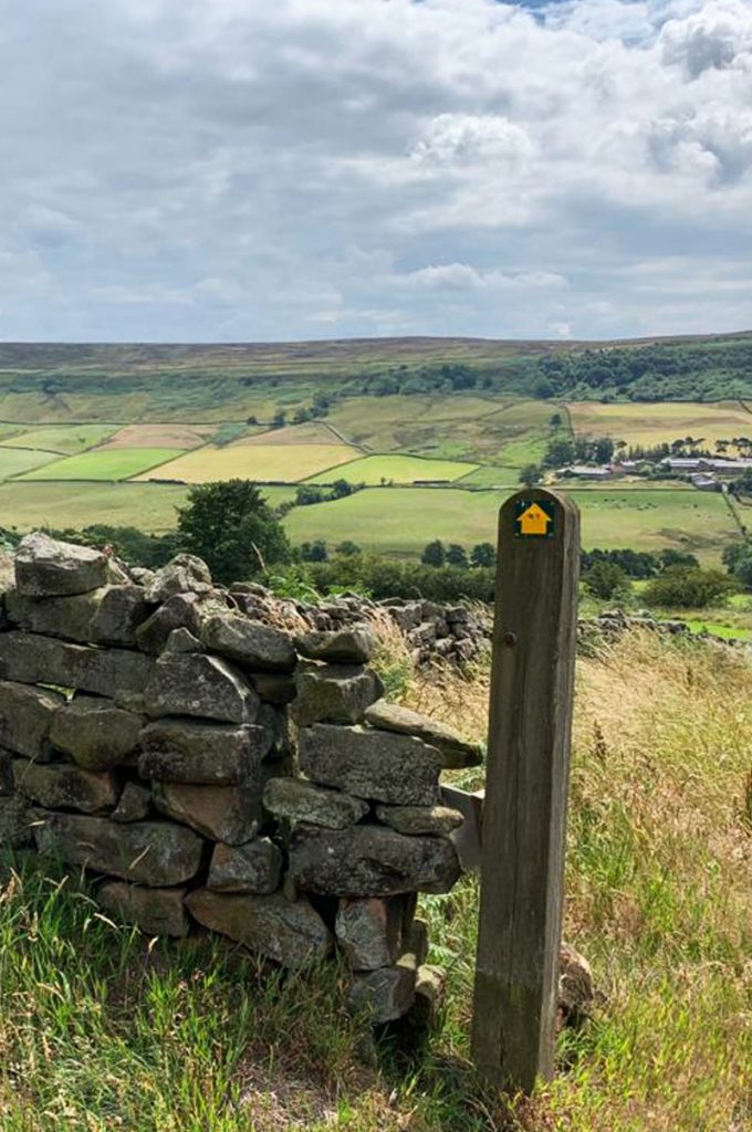 A rough stone wall next to a sign post with a yellow arrow on it pointing to a patch work of fields on a distant hill