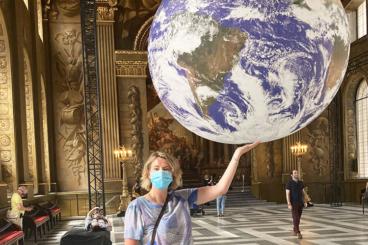 A blonde woman with a surgical face mask on her face standing in a large ornate hall pretending to balance a inflatable representation of the earth on her left hand