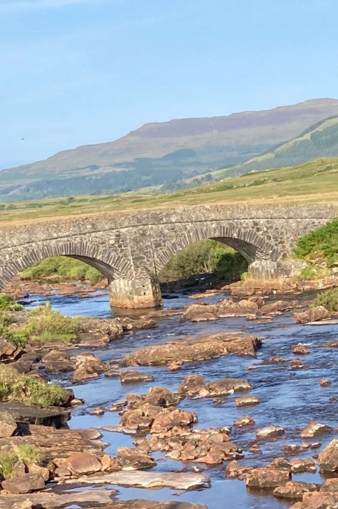A small stone bridge traverses a small babbling brooke in a picturesque highland setting.