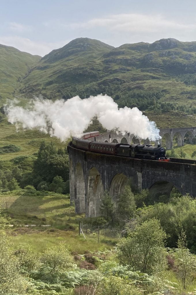 A steam train passes across a large stone viaduct in picturesque mountain scenary