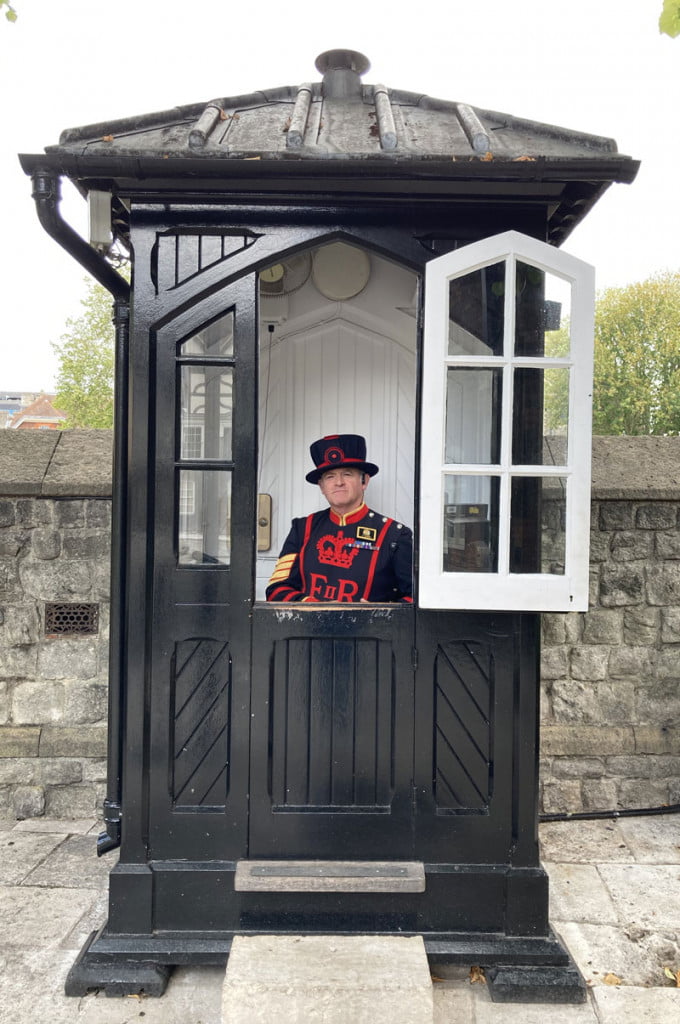 A Yeomen Warder in traditional black and red garb sat in a black sentry box with an open window at the Tower of London