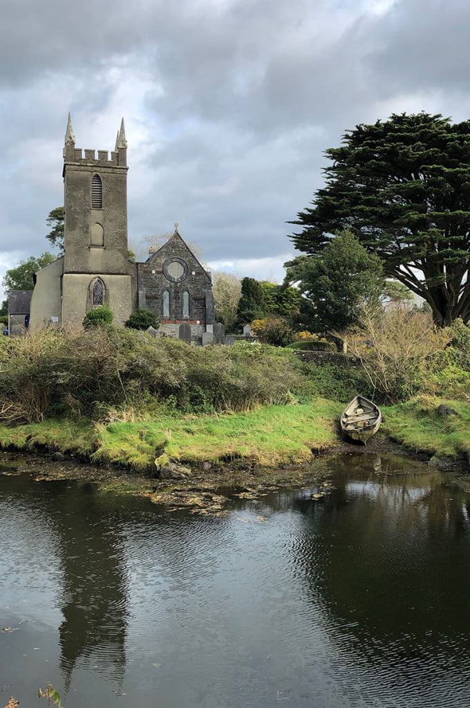 An old church nestles into the green countryside that surrounds a small river with a washed up rowing boat on the small river bank opposite