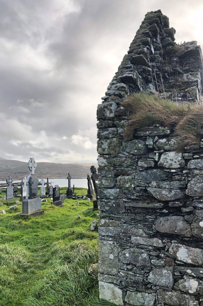 A remote Irish graveyard stands beyond a ruined stone hut on a bleak grassy hill overlooking a lake surrounded by distant mountains