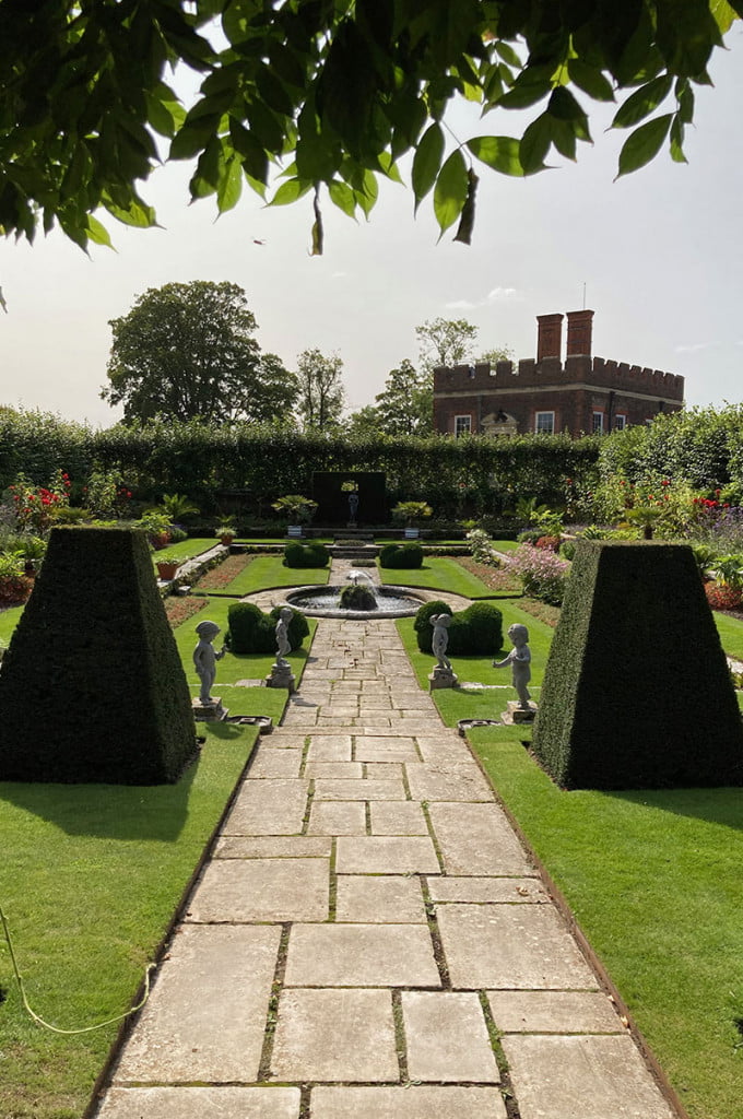 The manicured gardens of a stately home with triangular hedges and a geometric patterned path leading to a water feature in a circular pond