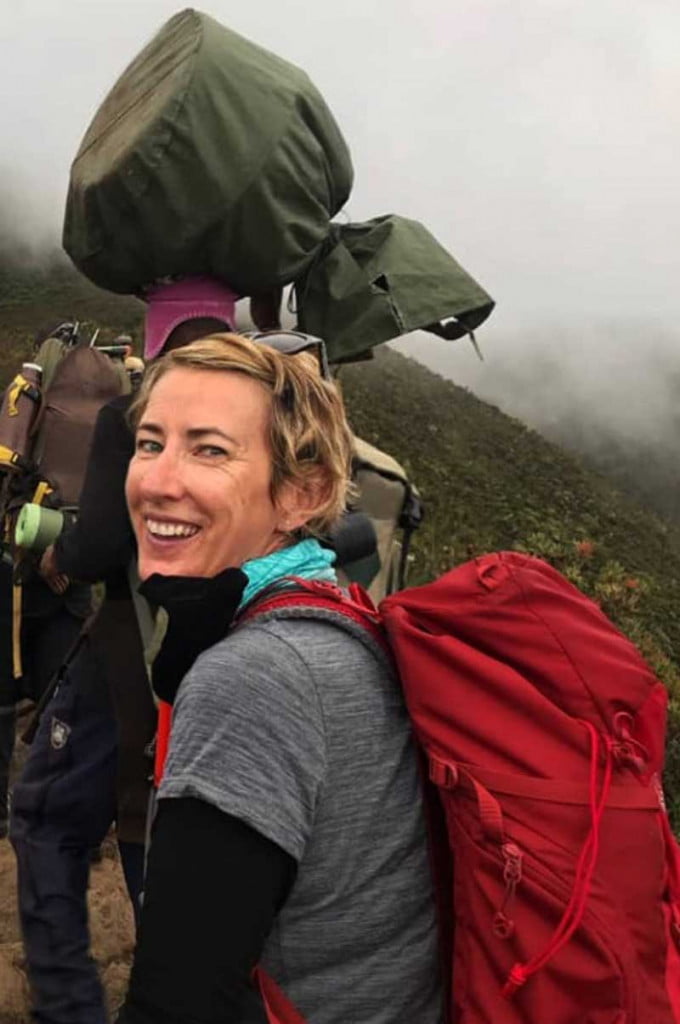 A blonde woman with a red rucksack turns to the camera to give a tired smile as she follows laden sherpas while Climbing Mount Kilimanjaro, 8 days on the Lemosho Route - Tanzania, Africa