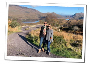 2 blonde women in sunglasses standing on a deserted path in front of a reflective lake and sprawling hills in The Ring of Kerry, Ireland