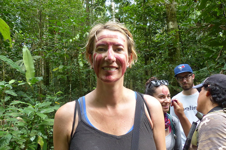 A blonde woman standing smiling in a jungle setting with red tribal paint all over her face