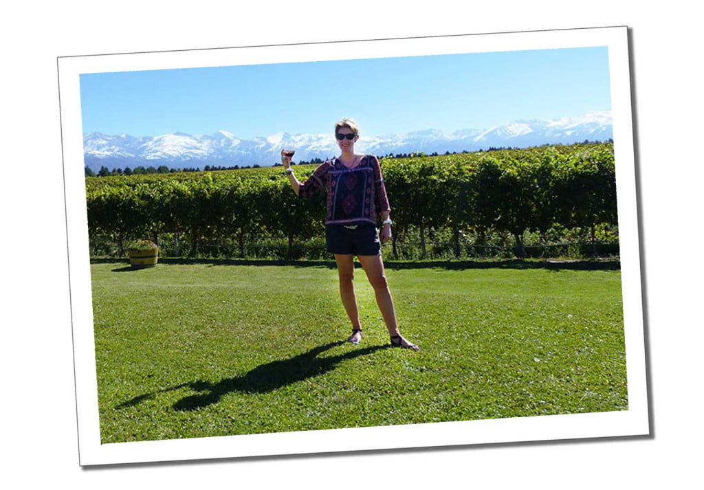 A Woman in sunglasses and purple top standing holding a glass of red wine in front of a large field of vineyards with a distant backdrop of snow capped mountains