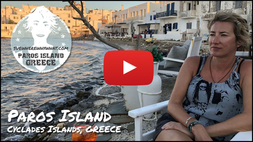 A blonde woman sitting on a white chair on the seas edge of a Greek Island resort with an orange cocktail with a large red YouTube button and white text overlay