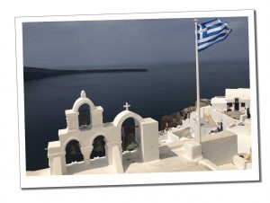 A greek flag flutters in the breeze next to a small white ornamental construction with 3 bells and a cross overlooking a dark sea in the bay beneath