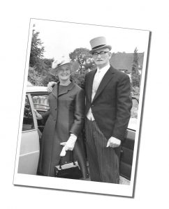 A black and white photo of a man and a woman in full dress outfits standing by the open door of a car