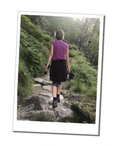 A Woman in a pink top and hiking boots walking away along a rough rocky path holding a water bottle in her right hand