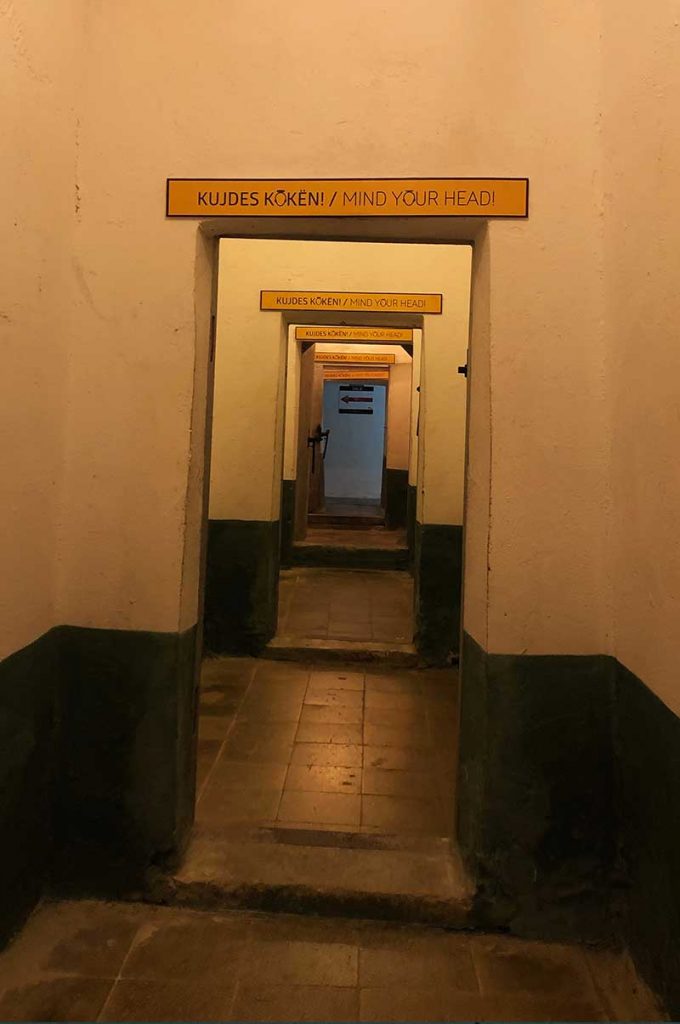 A dimly lit corridor leading through 5 small rooms to a blue wall in a drab an underground bunker with a mind your head sign above the open doorway of each