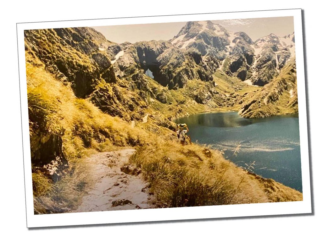 A person with a backpack on their head Hiking the Routeburn Track beside a deep blue lake