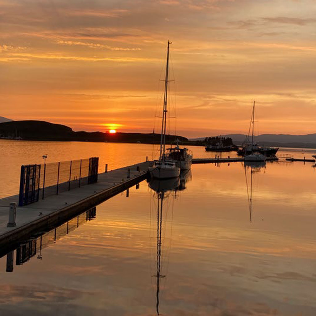 Silhouetted sailing boats moored by a jetty under an orange sunset sky over a tranquil calm loch