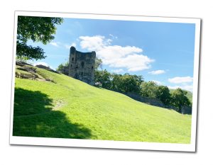 a ruined castle perched on a green sunny hillside with a park bench to it's left in the foreground