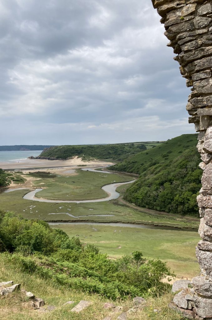 A ruined wall on a green hill leading to a river and ultimately the sea