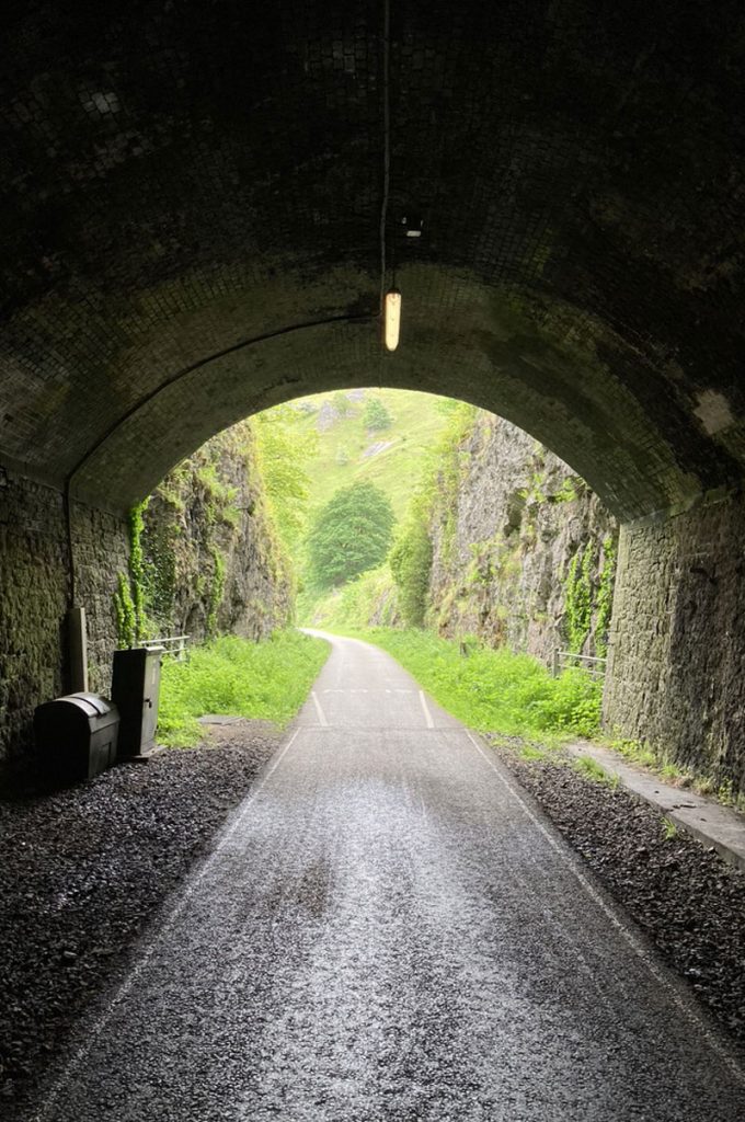 A view from inside a tunnel from a thin man made path winding through an English countryside valley featuring a small stone bridge and a dark tunnel through a green hillside