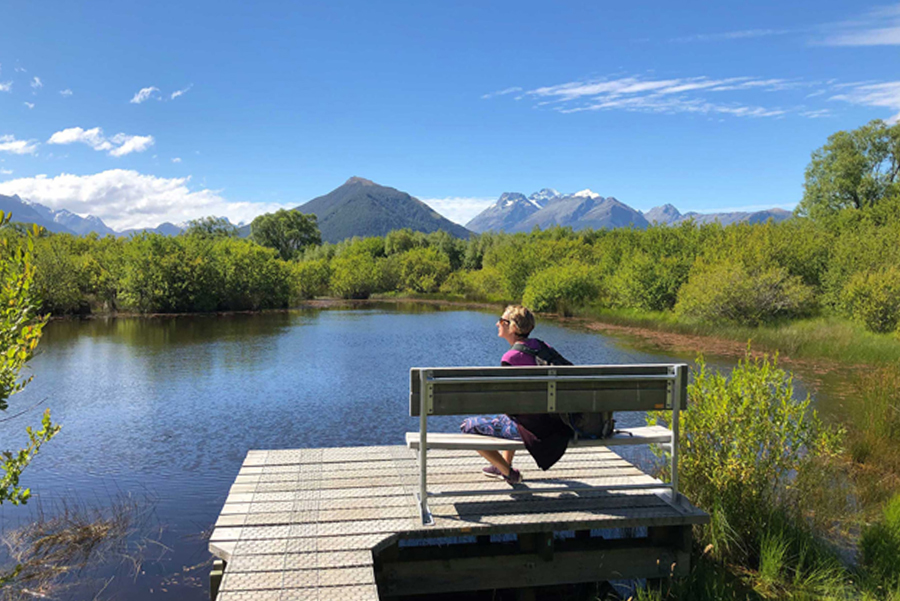 A blonde woman in sunglasses sitting on a park bench beside a still lake with a clear sky and distant mountain backdrop