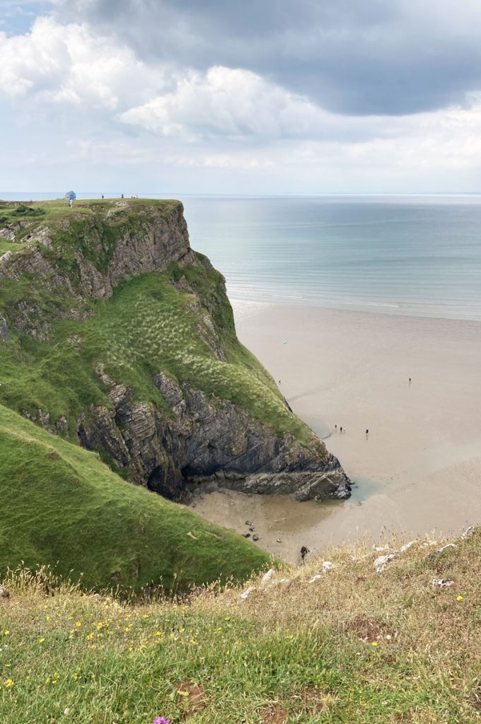 A view from a grassy hill of a cliff top over looking a calm Sandy bay with tiny ant like people walking on the sand