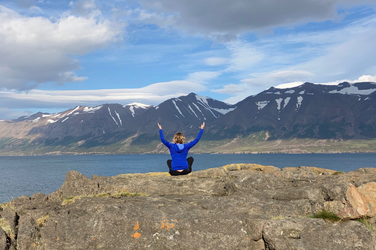 A Woman in a blue hooded top facing away towards snow capped mountains sitting on a large rock with her arms aloft