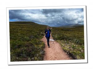 A woman in a blue coat and cap hiking along a dusty path across some flat green moorland
