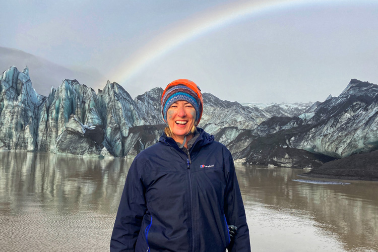 A Woman in a striped hat smiling in front of a backdrop of a tranquil glacier and rainbow