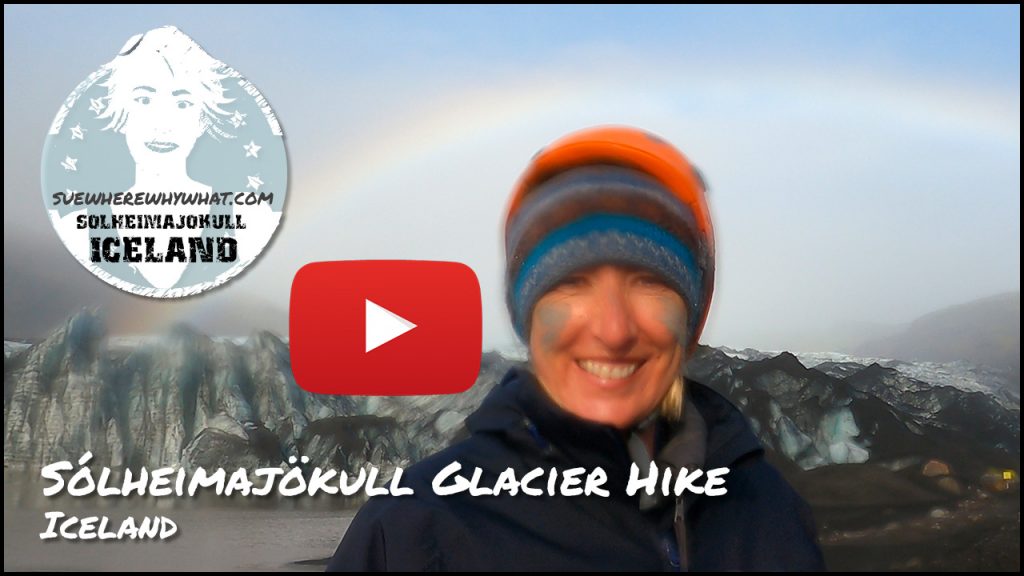 A Woman in a striped hat smiling in front of a backdrop of a tranquil glacier and rainbow with a large red youtube button in the foreground