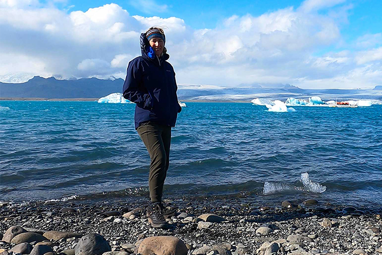 A Woman in blue coat and stripy hat stands on the shingle shore in front of a backdrop of White and grey glaciers floating on a turquoise stretch of flat water