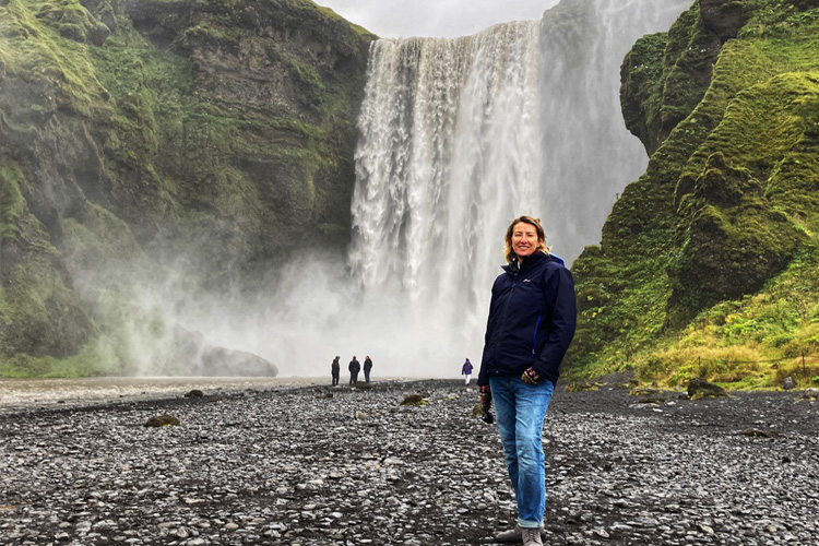 A Woman in jeans and blue jacket standing in front of a giant waterfall