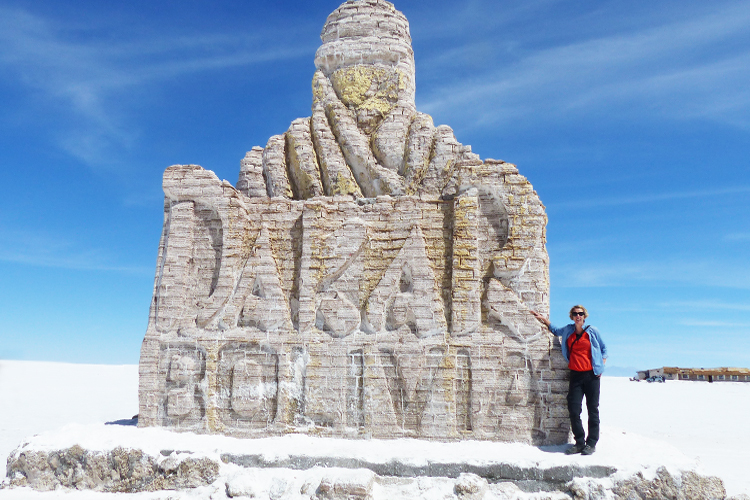 A Woman in a red top and sunglasses stannic in front of a large salt sculpture with the words DAKAR BOLIVIA carved on it.