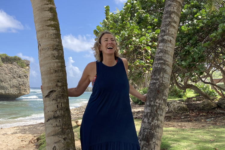 A blonde woman in a long blue dress standing laughing between two palm trees on a tropical beach