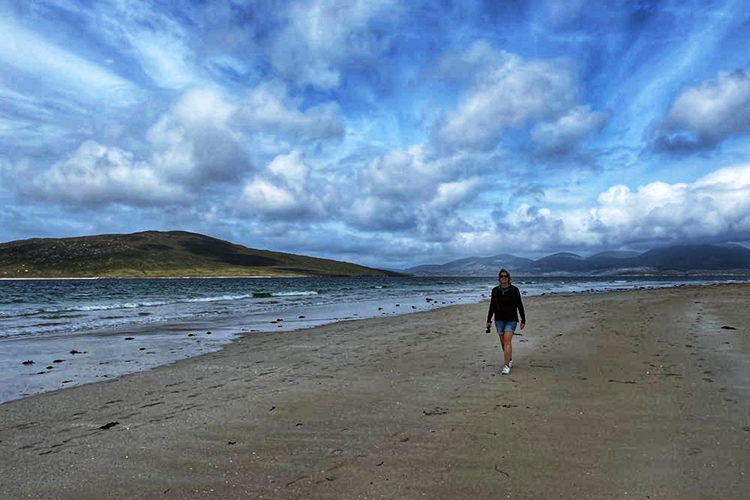 A Woman in a blue top walking along a deserted sandy beach with a dramatic skudding cloudy sky above