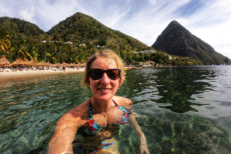 A Woman in Sunglasses standing in a clear tropical sea with a mountainous backdrop