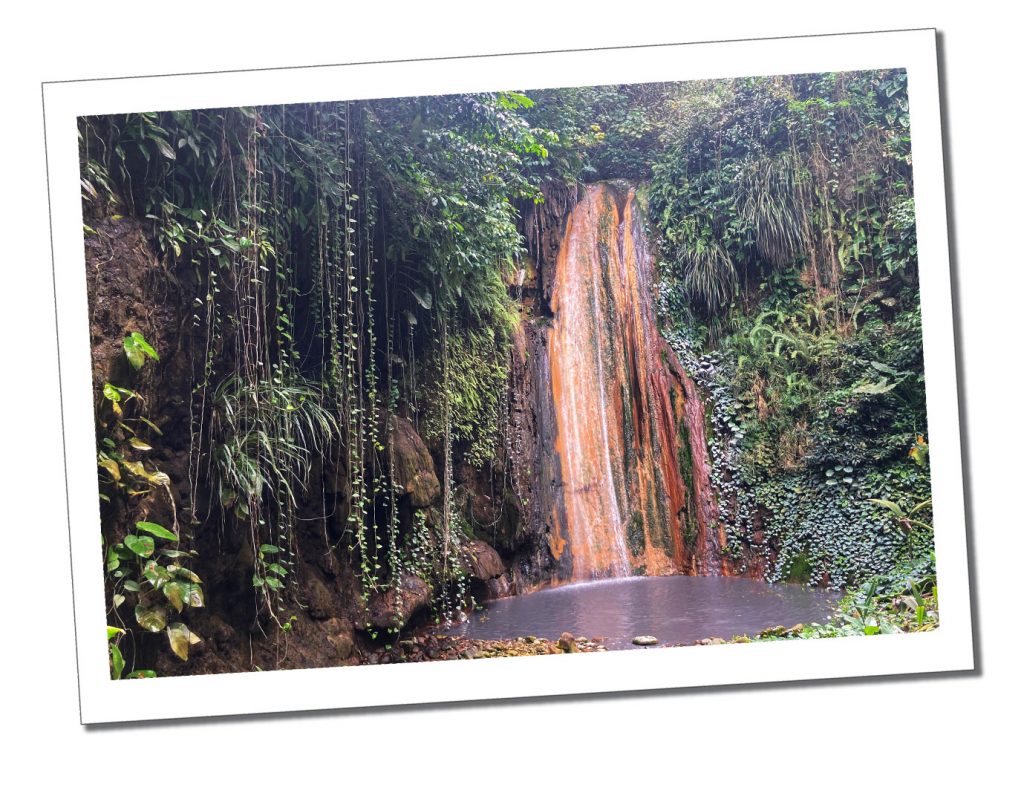 A Golden coloured rock face behind a waterfall in a tropical setting