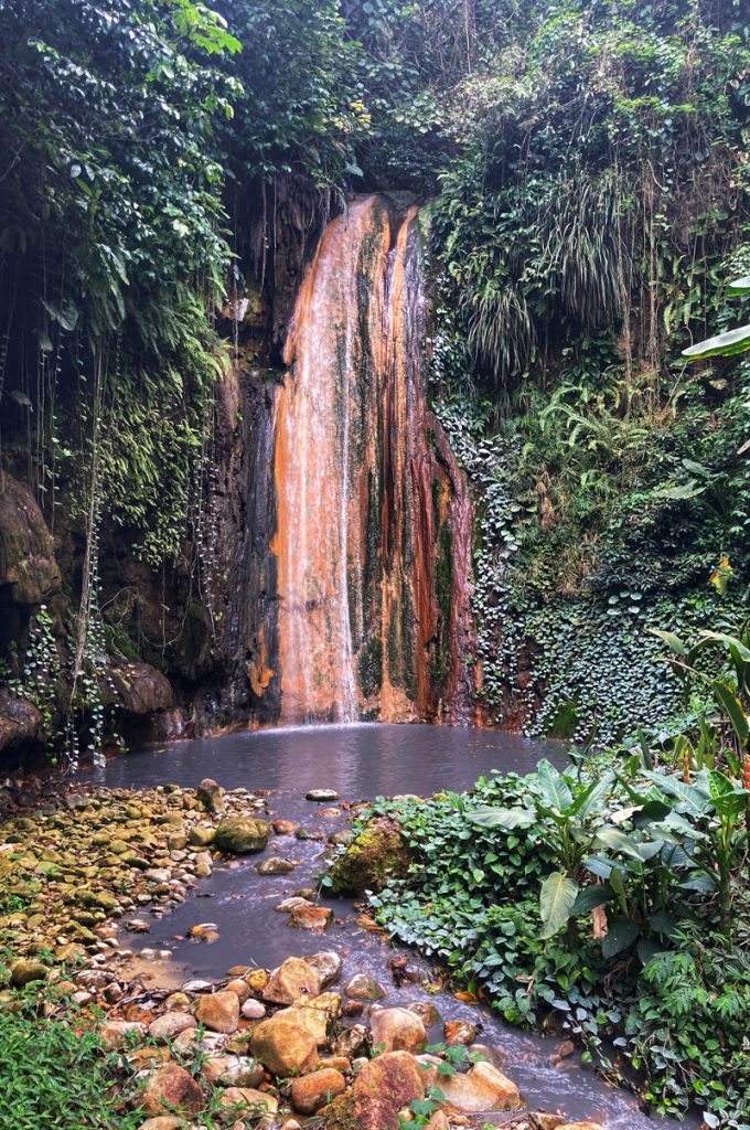 A Golden coloured rock face behind a waterfall in a tropical setting