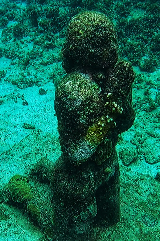 A sculpture depicting stone person praying on it's knees in an underwater scene in Grenada.