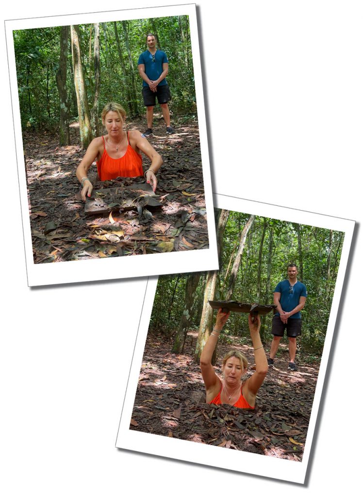 A woman in an orange top grimaces as she holds a flat board above her head while standing in hole in a leafy clearing watched by a man in shorts and a blue top