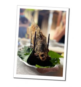A deep fried fish facing the camera on a stand on a plate of leaves