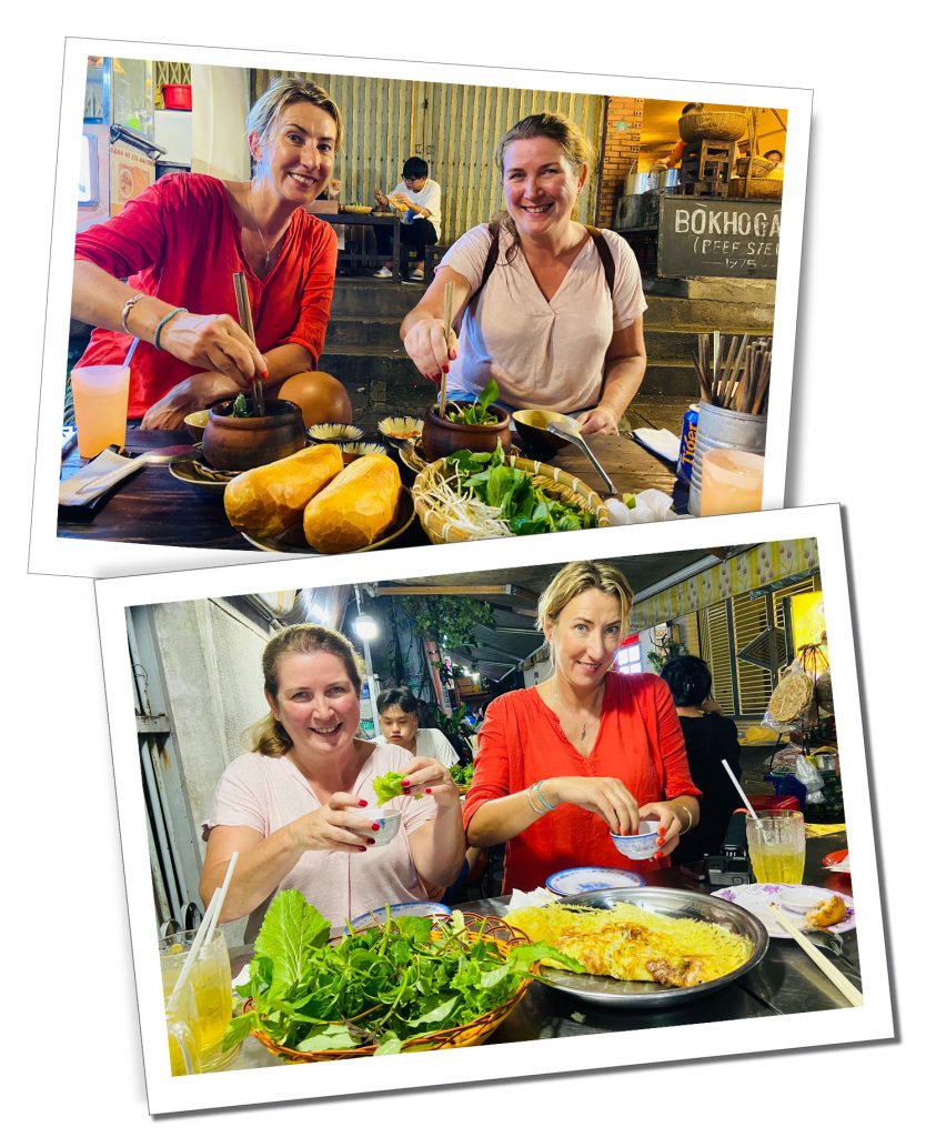 Two woman sit smiling eating Vietnamese street food at night
