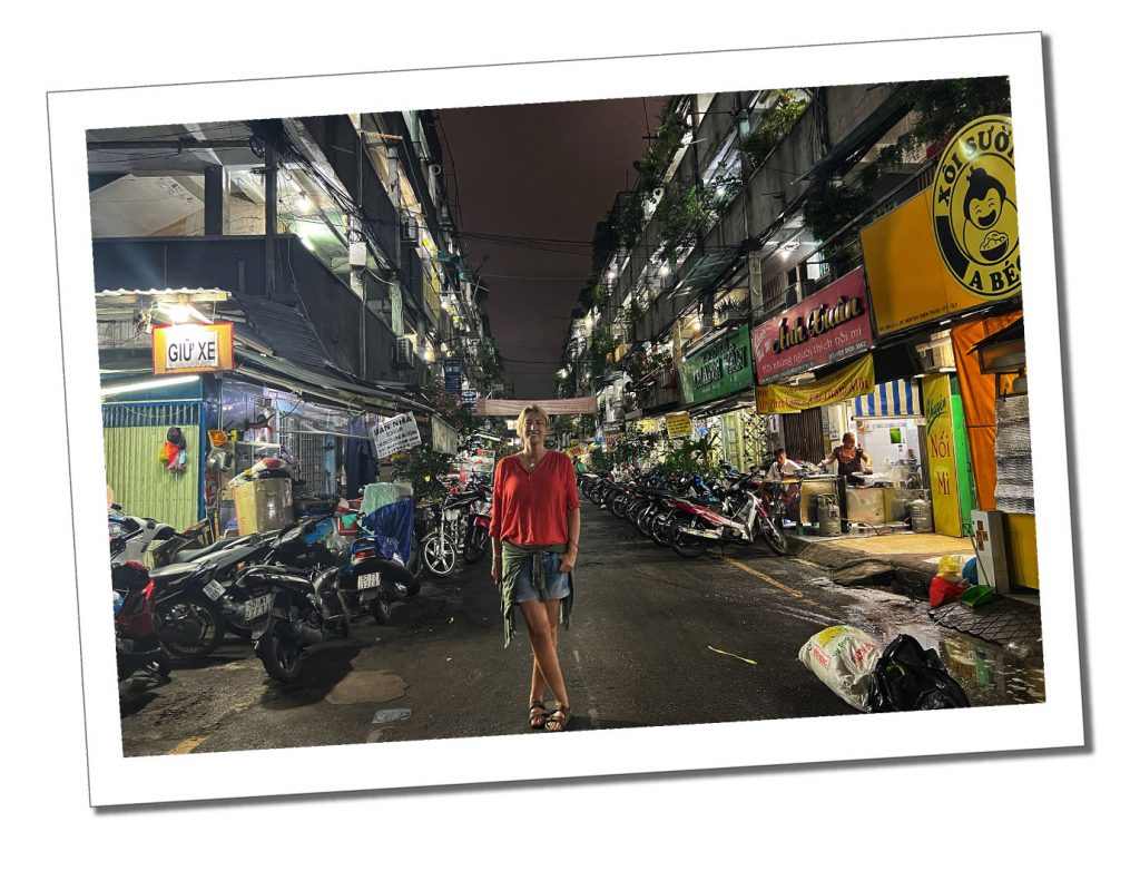 A Woman in an Orange top and denim shorts smiling at the camera standing in a brightly lit Vietnamese street full of motor bikes and scooters