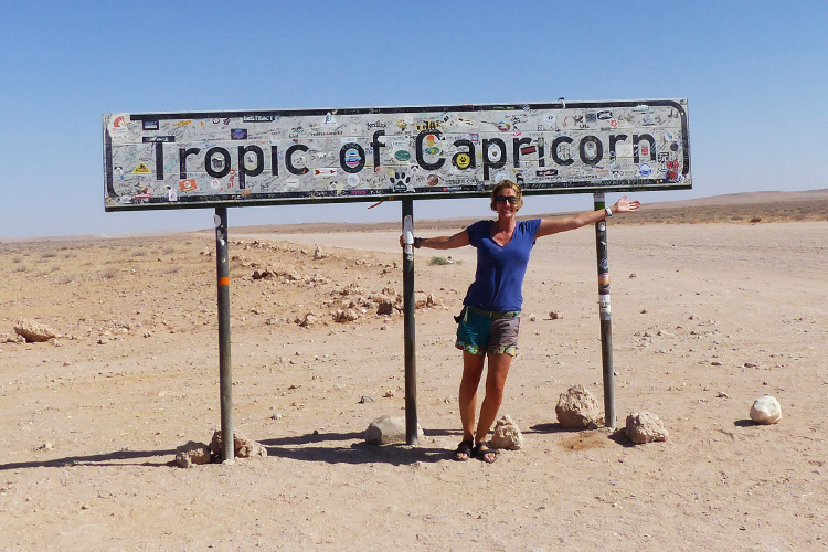 A Woman in a blue top leaning against a sign which reads the tropic of capricorn