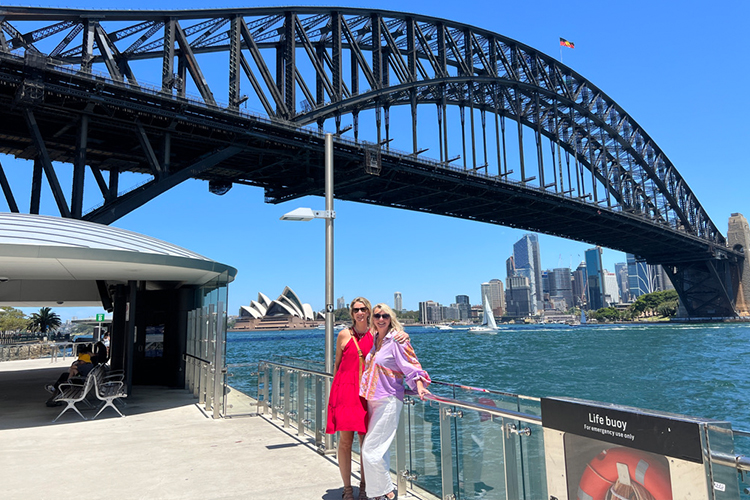 Two blonde women standing leaning against a metal railing underneath the Sydney Opera House in Australia