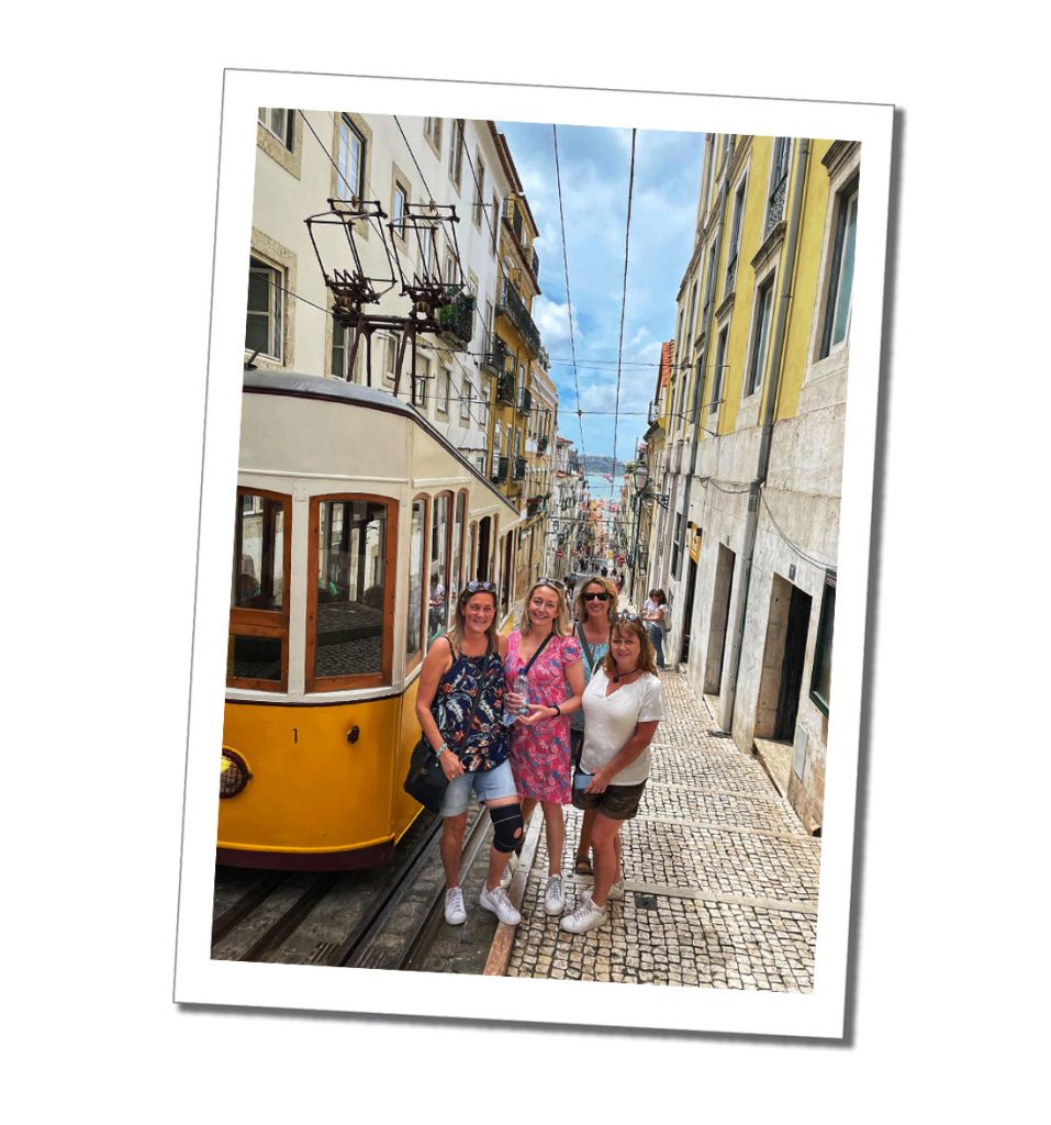 4 women standing next to a yellow tram car at the top of a hill in a narrow street