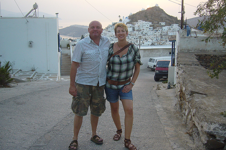 A man and a woman standing arm in arm looking at the camera with a backdrop of white houses and a mountain